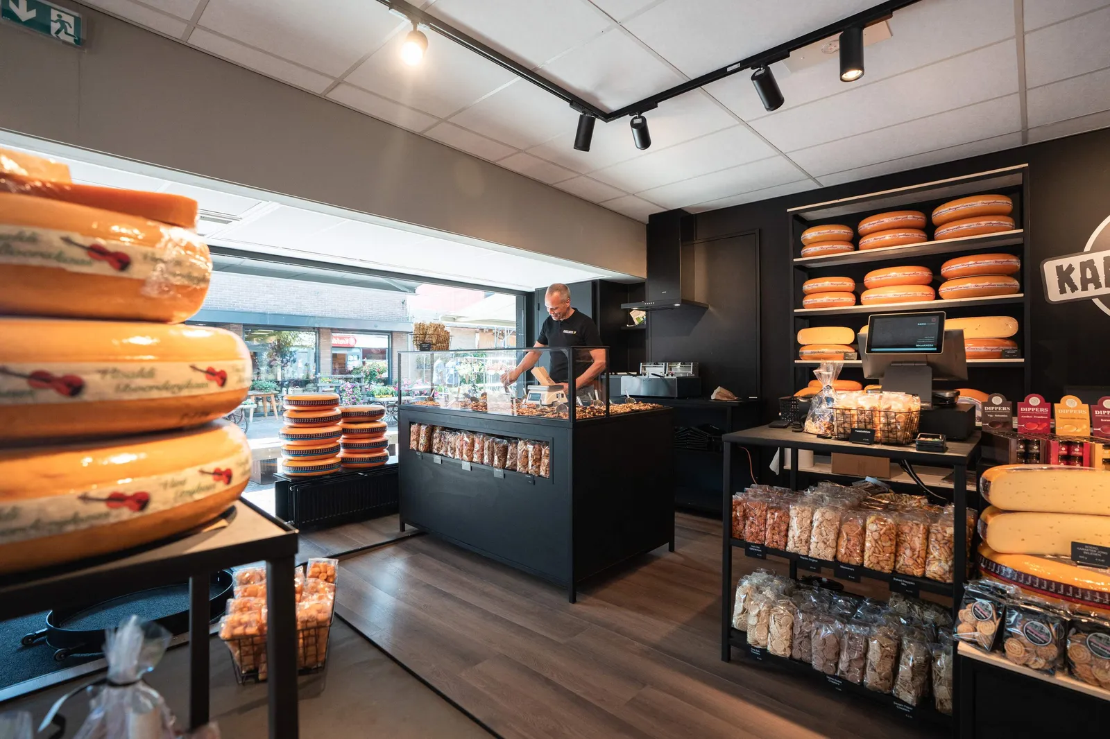 Binnenkant van Kaasloods 28: kaaswielen op de schappen, noten en Rik achter de toonbank
